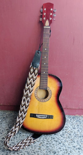 Brown patterned Wayuu guitar strap on an acoustic guitar resting against a maroon wall. 